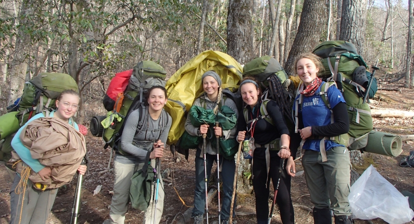 A group of people wearing large backpacks smile while standing in a wooded area. 